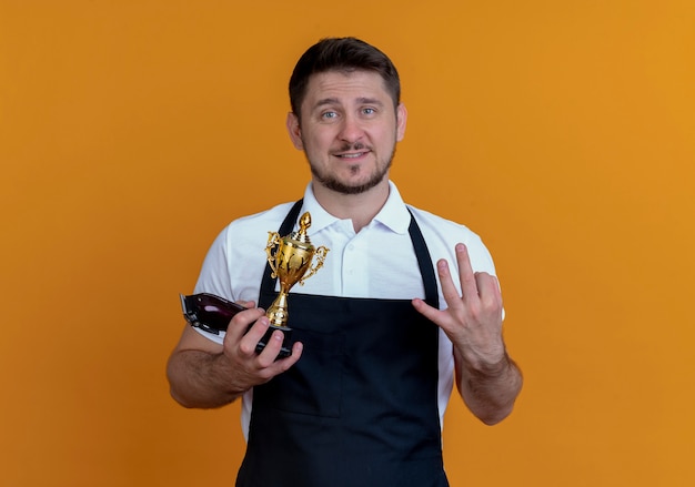 barber man in apron holding beard trimmer and trophy  showing number three smiling standing over orange wall