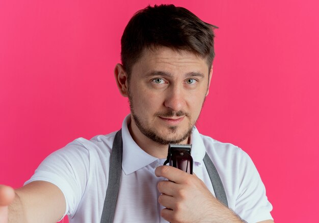barber man in apron holding beard trimmer  with smile on face standing over pink wall