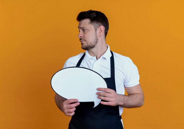 barber man in apron holding blank speech bubble sign looking aside displeased standing over orange wall