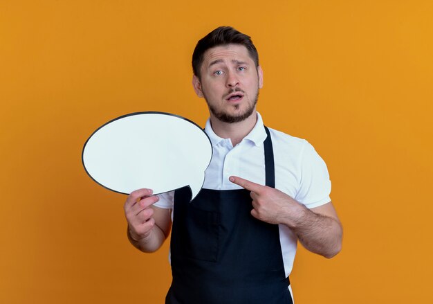 barber man in apron holding blank speech bubble sign pointign with finger to it with confuse expression standing over orange wall