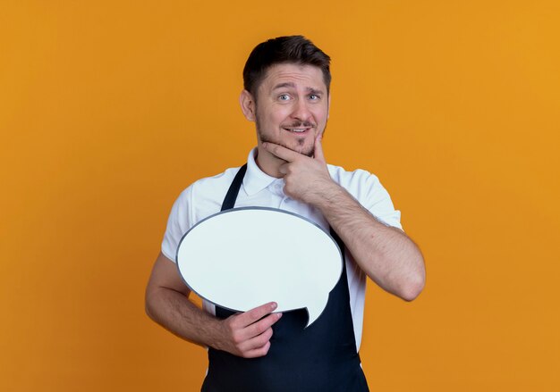 barber man in apron holding blank speech bubble sign  with hand on ching smiling and thinking standing over orange wall