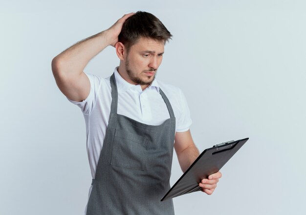 Barber man in apron holding clipboard looking at it confused scratching his head standing over white background