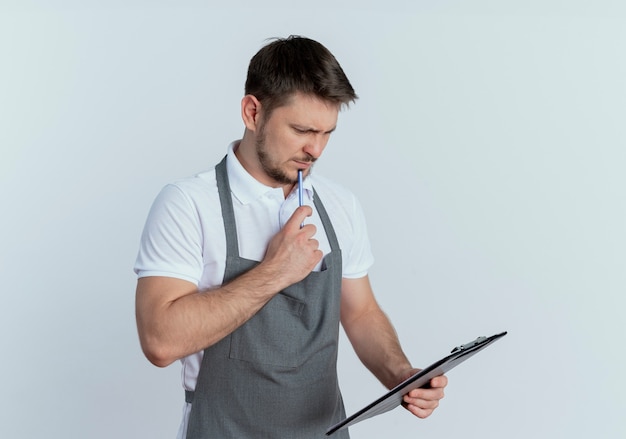 barber man in apron holding clipboard looking at it with pensive expression on face standing over white wall