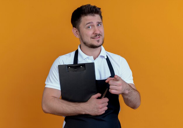 barber man in apron holding clipboard pointing with finger smiling confident standing over orange wall