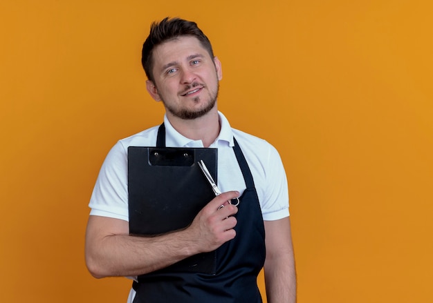 Barber man in apron holding clipboard and scissors looking at camera with smile on face standing over orange background