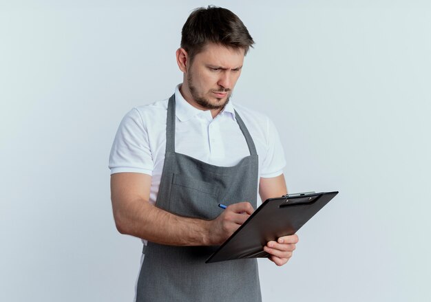 barber man in apron holding clipboard writing something with serious face standing over white wall