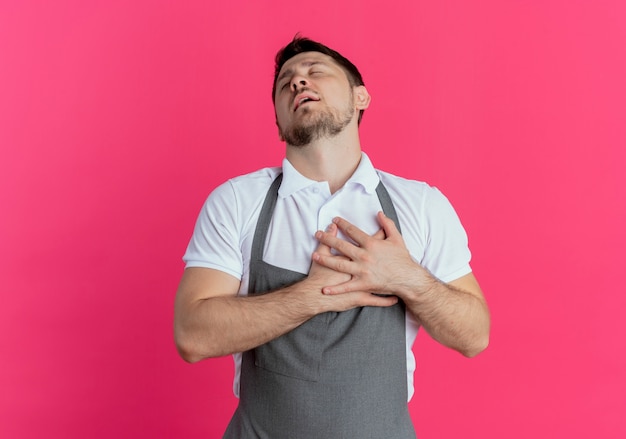 barber man in apron holding crossed arms on his chest feeling thankful with closed eyes standing over pink wall