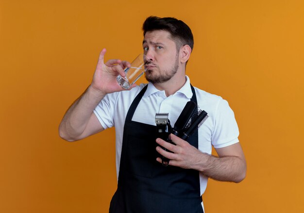 Barber man in apron holding hair brush and beard trimmer drinking water looking at camera with skeptic expression standing over orange background