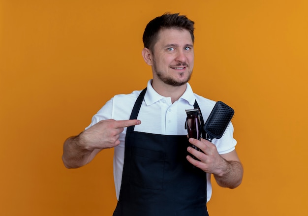 Barber man in apron holding hair brush and beard trimmer pointing with finger at them smiling confident standing over orange background