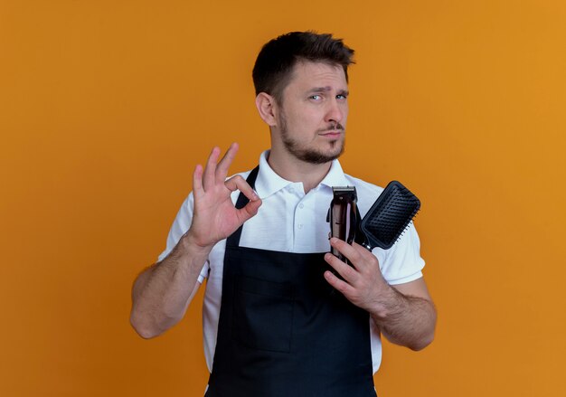 barber man in apron holding hair brush and beard trimmer showing ok sign looking confident standing over orange wall