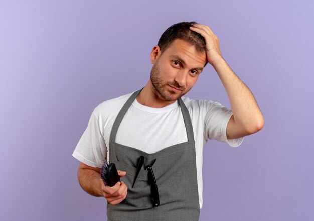 Barber man in apron holding hair brush looking to the front with confident expression standing over purple wall