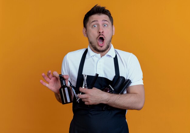 Barber man in apron holding hair brushes , spray and scissors looking at camera surprised standing over orange background