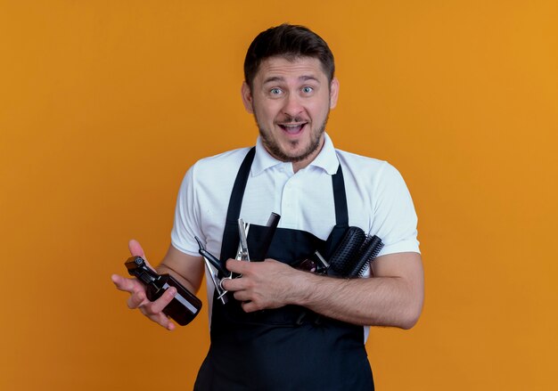 barber man in apron holding hair brushes , spray and scissors  smiling standing over orange wall