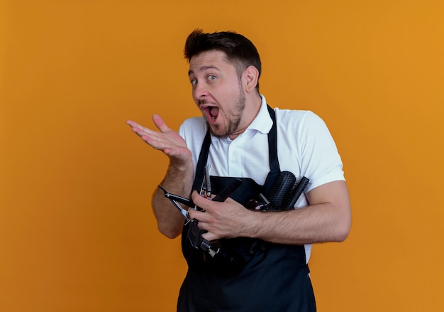 barber man in apron holding hair brushes , spray and scissors  with happy face surprised standing over orange wall