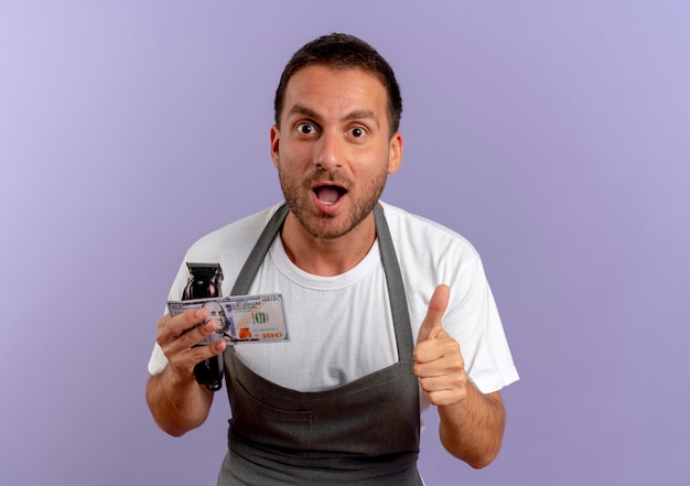 Barber man in apron holding hair cutting machine and cash happy and excited showing thumbs up standing over purple wall