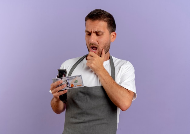 Barber man in apron holding hair cutting machine and cash loking amazed and surprised standing over purple wall