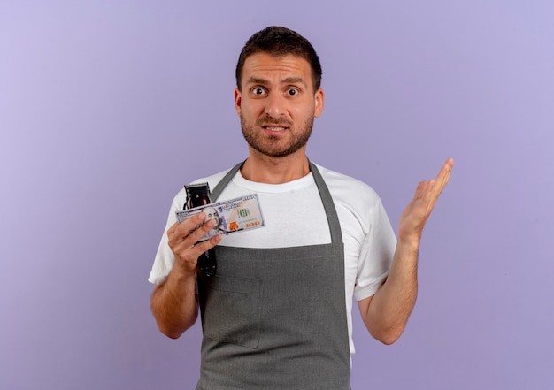 Barber man in apron holding hair cutting machine and cash looking to the front confused standing over purple wall