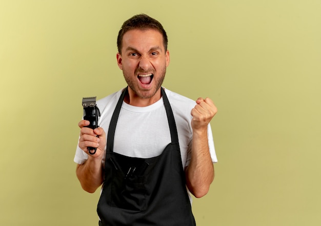 Barber man in apron holding hair cutting machine clenching fist happy and excited standing over light wall