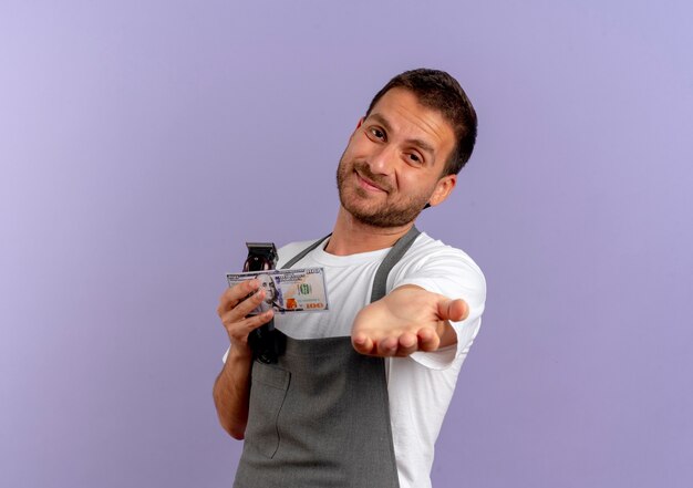 Barber man in apron holding hair cutting machine holding hand in front of himself asking for money standing over purple wall