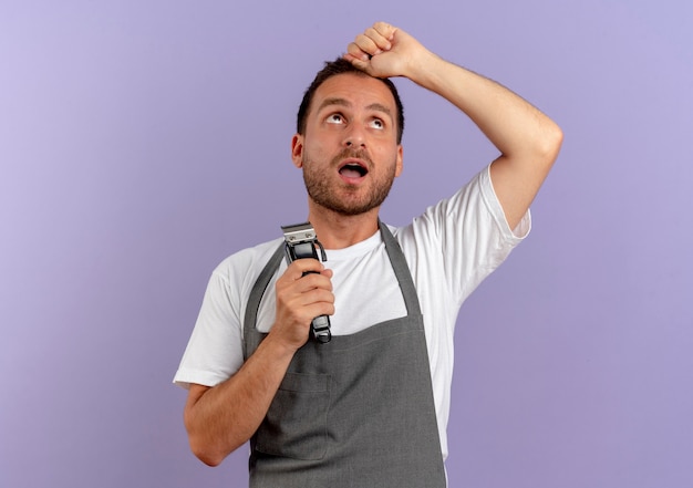 Free photo barber man in apron holding hair cutting machine looking up with confuse expression standing over purple wall