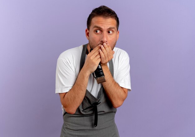 Barber man in apron holding hair cutting machine shocked covering mouth with hands standing over purple wall