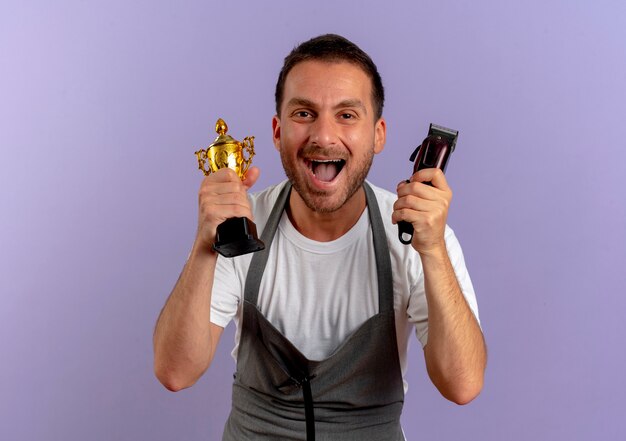 Barber man in apron holding hair cutting machine and trophy happy and excited smiling cheerfully standing over purple wall