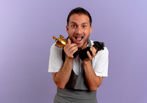 Barber man in apron holding hair cutting machine and trophy looking to the front happy and excited standing over purple wall