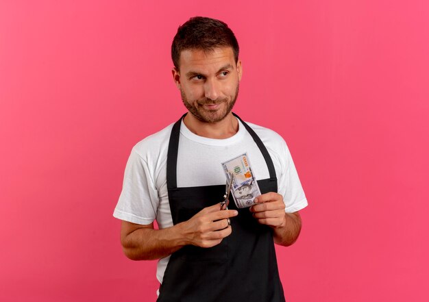 Barber man in apron holding scissors and cash looking aside with skeptic expression standing over pink wall