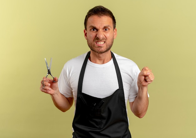 Foto gratuita uomo del barbiere in grembiule che tiene le forbici stringendo il pugno guardando in avanti con la faccia arrabbiata in piedi sopra la parete chiara