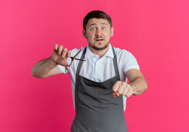 Barber man in apron holding scissors clenching fist looking worried standing over pink background