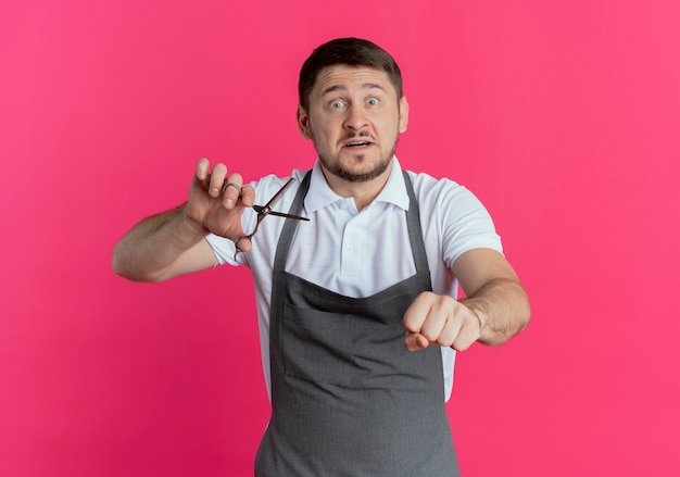 barber man in apron holding scissors clenching fist looking worried standing over pink wall