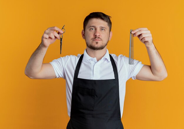 Barber man in apron holding scissors and comb looking at camera with serious face standing over orange background