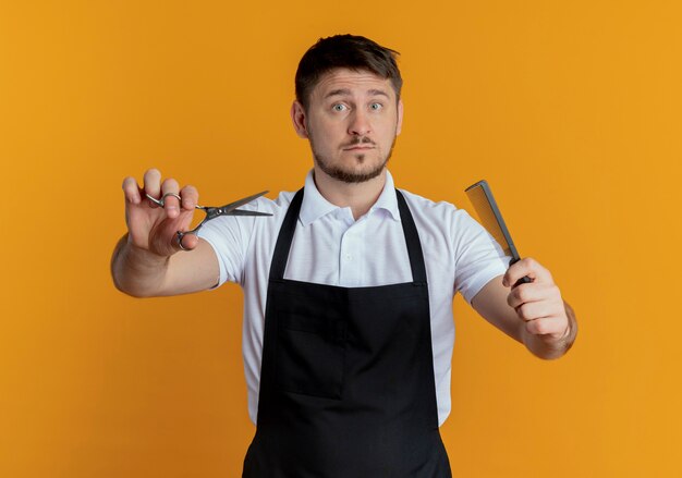 Barber man in apron holding scissors and comb looking at camera with serious face standing over orange background