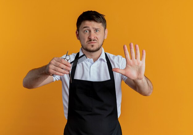 Barber man in apron holding scissors and comb making stop sing with open hand looking at camera with serious face standing over orange background