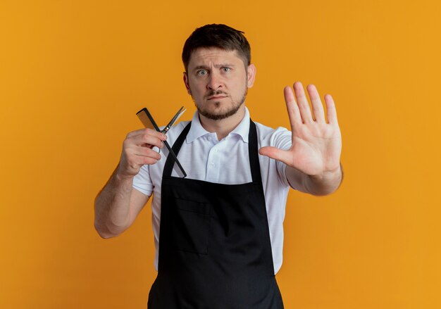 Barber man in apron holding scissors and comb making stop sing with open hand looking at camera with serious face standing over orange background