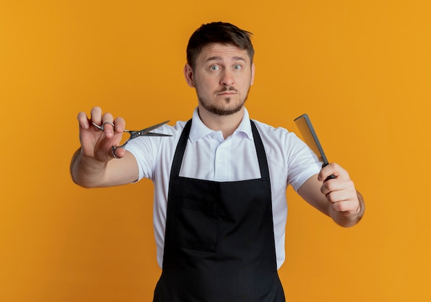 barber man in apron holding scissors and comb  with serious face standing over orange wall