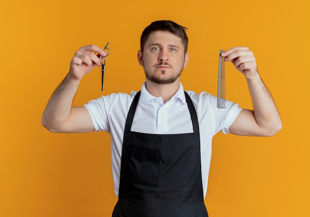Free photo barber man in apron holding scissors and comb  with serious face standing over orange wall