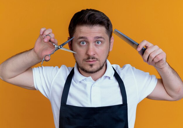 barber man in apron holding scissors and comb  with wide ope eyes standing over orange wall