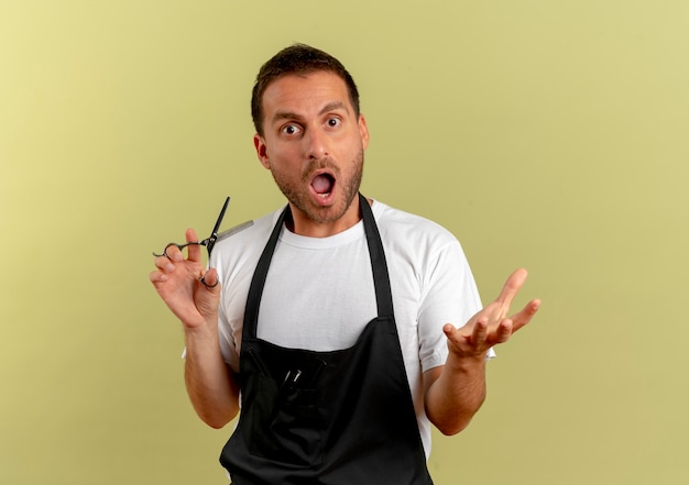 Barber man in apron holding scissors looking to the front amazed and surprised standing over light wall