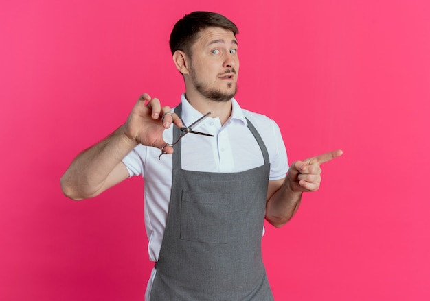 barber man in apron holding scissors pointing with index figer to the side looking confident standing over pink wall