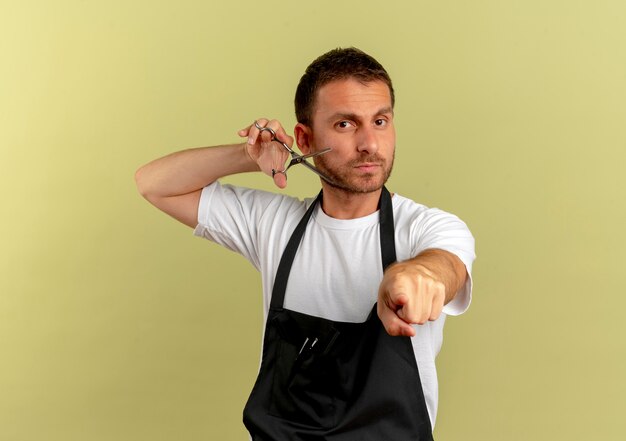 Barber man in apron holding scissors pointing with index finger to the front with confident expression standing over light wall