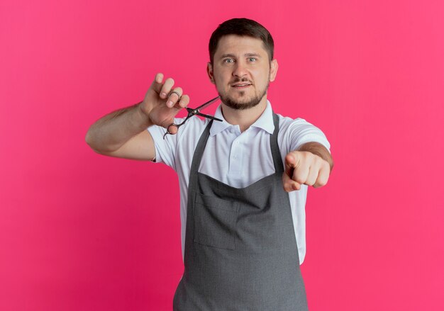 barber man in apron holding scissors pointing with index finger standing over pink wall