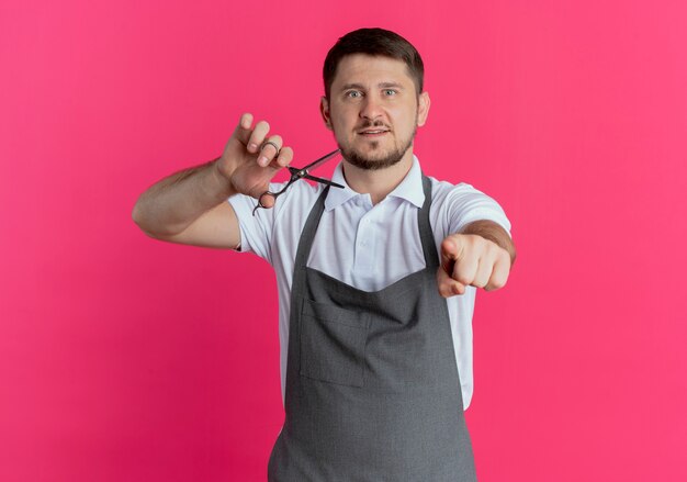 Barber man in apron holding scissors pointing with indexinger to camera standing over pink background