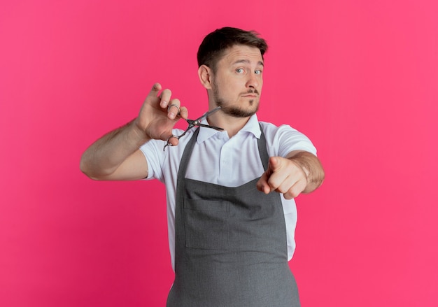 Barber man in apron holding scissors pointing with indexinger to camera with confident expression standing over pink background