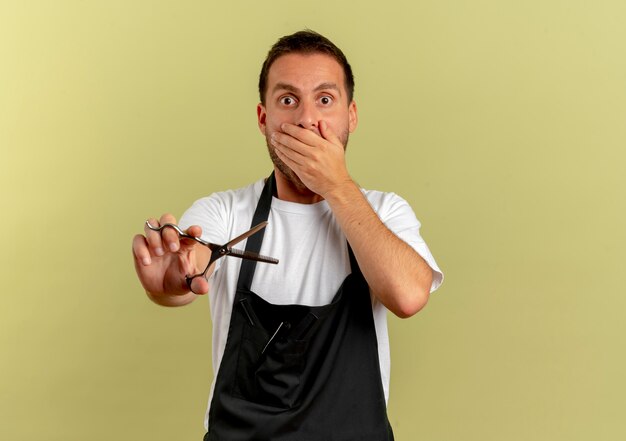 Barber man in apron holding scissors shocked covering mouth with hand standing over light wall