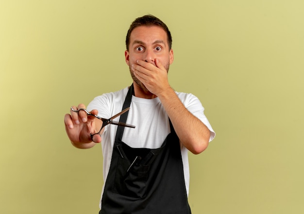 Barber man in apron holding scissors shocked covering mouth with hand standing over light wall