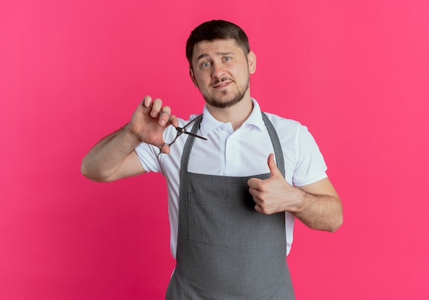 Foto gratuita barbiere uomo in grembiule tenendo le forbici che mostra i pollici in su con il sorriso sul viso in piedi sopra il muro rosa