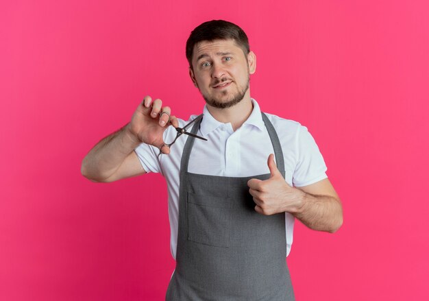 barber man in apron holding scissors showing thumbs up  with smile on face standing over pink wall