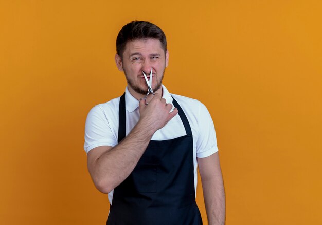 Barber man in apron holding scissors trying to cut his nose standing over orange background