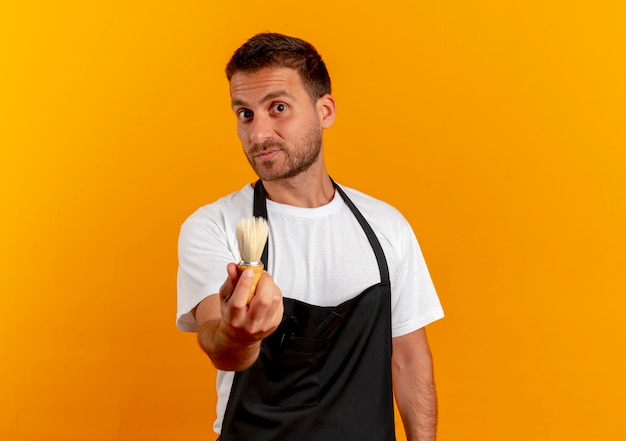 Barber man in apron holding shaving brush looking to the front with confident expression standing over orange wall